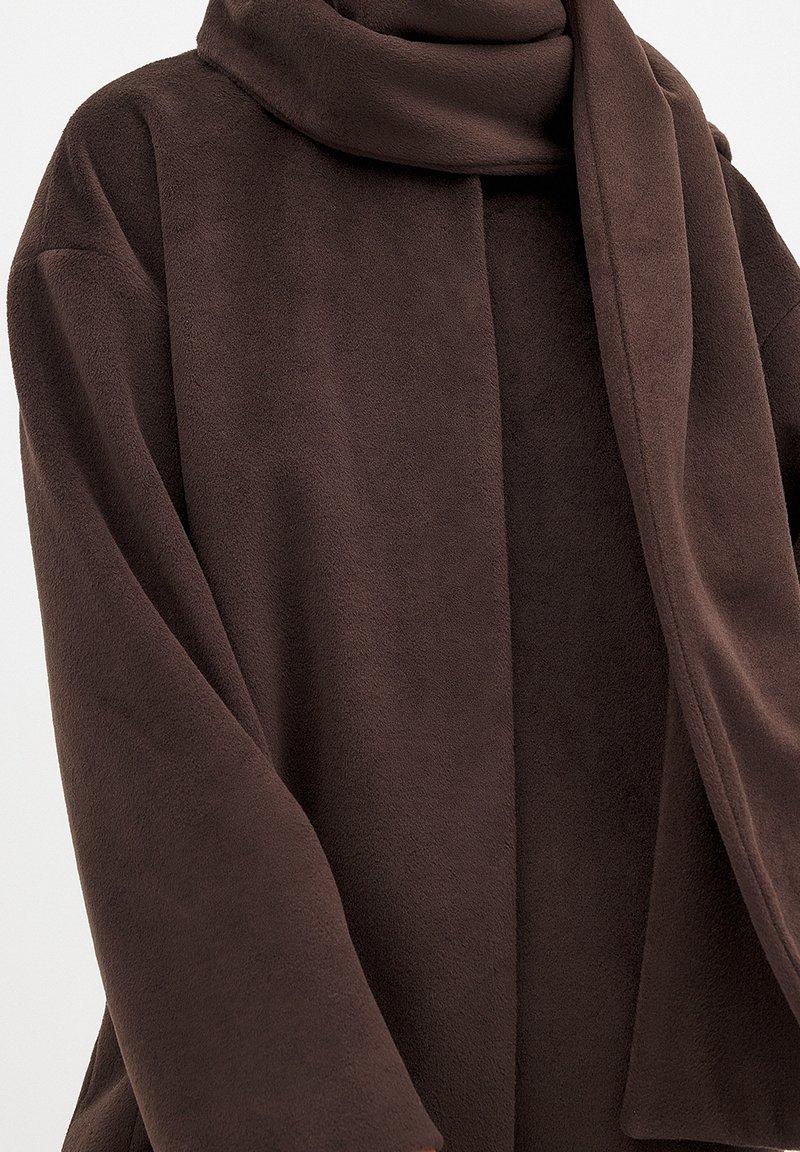 Abrigo de forro polar marrón con una textura suave, que presenta un cuello ancho y una abertura frontal. Las mangas son holgadas, proporcionando comodidad y calidez.