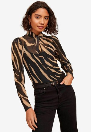 Blusa trasparente a maniche lunghe con motivo a strisce di tigre in beige e nero, colletto alto con fiocco, abbinata a jeans neri e una cintura.