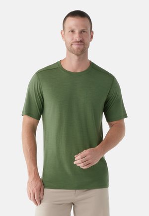Uomo con capelli corti che indossa una maglietta verde a maniche corte con scollo rotondo e pantaloni beige, in piedi contro uno sfondo bianco uniforme.