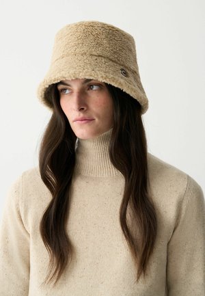 Beige Bucket Hat aus weichem Stoff, mit einer strukturierten Oberfläche und einem kleinen Logodetail an der Seite. Trägt man zu einem hellbeigen Pullover.