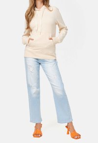 Felpa beige con cappuccio e tasca frontale, abbinata a jeans chiari strappati e sandali con tacco a frange arancioni.