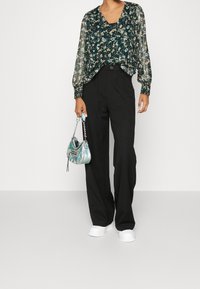 Femme portant un chemisier transparent vert à motifs floraux, un pantalon noir à jambes larges, des chaussures blanches, tenant un petit sac à main métallique argenté par la lanière.