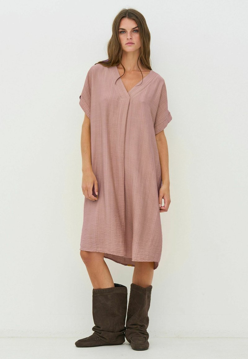Femme aux longs cheveux bruns portant une robe ample rose poudré longueur genou et des bottes marron foncé décontractées sur un fond blanc uni.