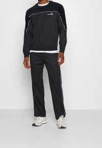 Uomo vestito con una tuta nera a fantasia con piping bianco e sneakers bianche, in piedi su uno sfondo plain chiaro.