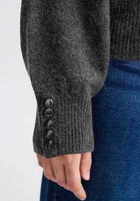 Manica di un maglione lavorato a maglia grigio scuro con polsino a coste e cinque bottoni neri lucidi. Texture morbida con fibre visibili.