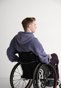 Lilla hettegenser, mørke bukser, sitter i en svart rullestol med stilige hjul og ergonomisk ryggstøtte, minimal bakgrunn.
