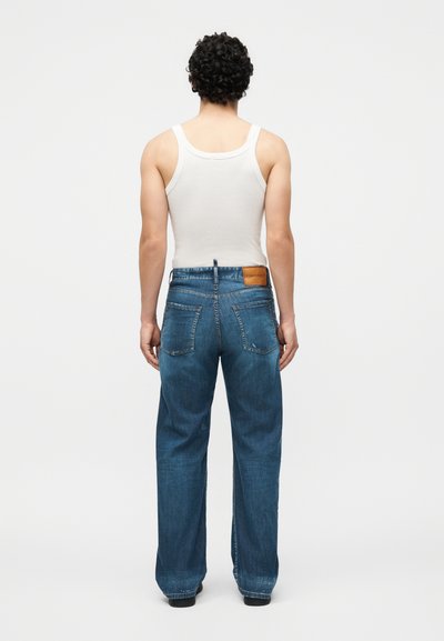 Άνδρας με σγουρά μαλλιά που φοράει λευκό αμάνικο μπλουζάκι και άνετο μπλε τζιν, στέκεται γυρισμένος πλάτη σε απλό λευκό φόντο.