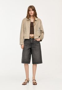Giubbotto bomber in suede beige con dettagli borchiati, abbinato a pantaloni culotte in denim scuro e sandali marroni con cinturini.
