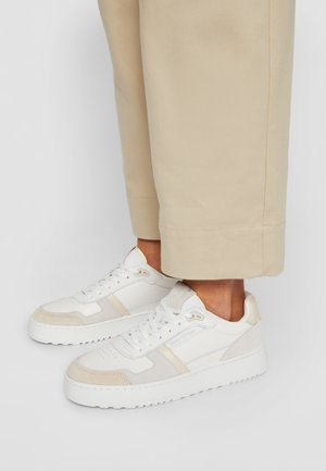 Hvide lave sneakers med beige accenter båret med beige korte bukser på en person, der står mod en ensfarvet baggrund.