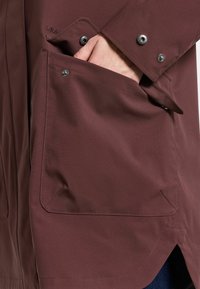 Bordeaux waterdichte jas met een grote zak, drukknoopsluitingen, een soepele textuur en een afgeronde zoom. Subtiele branding zichtbaar op de stof.