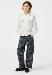 Blusa bianca con colletto arricciato, davanti con bottoni; abbinata a jeans scuri con stampa di unicorni. Scarpe nere completano il look.