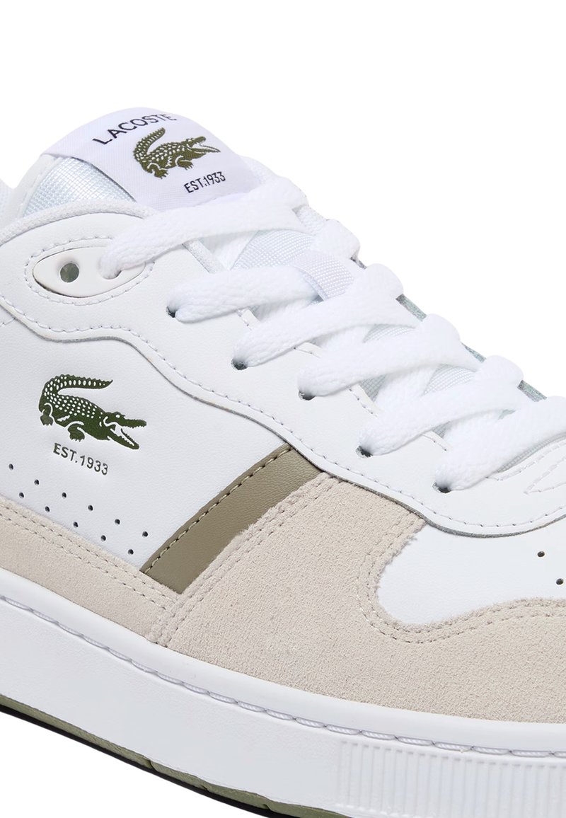 Lacoste-sneakers har en hvid læder-overdel med beige ruskindsdetaljer, tekstureret gummisål og en grøn krokodille-logo.