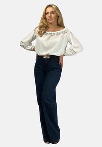 Biała bluzka z koronkowymi akcentami przy dekolcie i rękawach, zestawiona z ciemnoniebieskimi spodniami dzwonami. Modelka nosi czarny pasek.