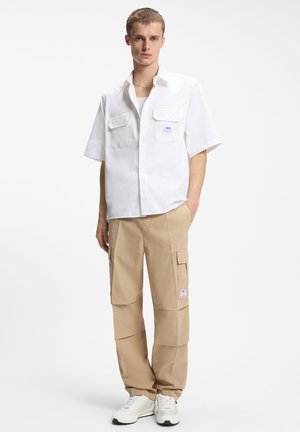 Jeune homme portant une chemise blanche à manches courtes boutonnée, un pantalon cargo beige et des baskets blanches, debout avec une main dans la poche.