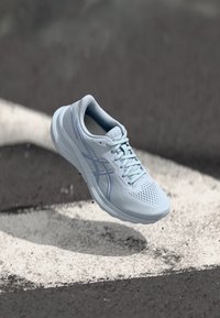 Scarpa da corsa azzurro chiaro con tomaia in rete traspirante, forma liscia e arrotondata, suola testurizzata e dettagli blu sul logo laterale.
