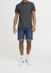 Muž v tmavě šedém polo tričku s červeným lemem, srolovaných džínových šortkách, bílých ponožkách a bílých teniskách s černými pruhy.