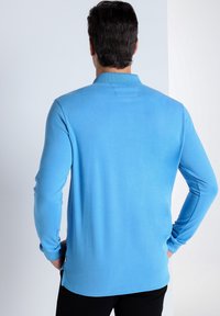 Langärmliges Polohemd in Hellblau, aus weichem Stoff gefertigt. Verfügt über einen gerippten Kragen und ein kleines Rückentag, mit seitlichen Schlitzen am Saum.