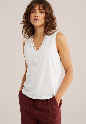 Vrouw draagt een mouwloos wit shirt met kanten details bij de V-hals en een bordeauxrode broek, staand met handen in de zakken tegen een effen achtergrond.