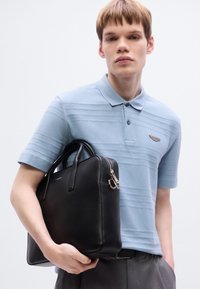 Μαύρη δερμάτινη τσάντα με χειρολαβές στην κορυφή και ασημένιο μεταλλικό εξάρτημα, σε συνδυασμό με μπλε ανοιχτή πόλο μπλούζα με υφασμένους κάθετους ρίγες και λογότυπο.