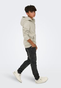 Un garçon aux cheveux bruns portant un sweat à capuche gris clair, un jean noir et des baskets blanches, marchant avec une main dans la poche sur fond uni.