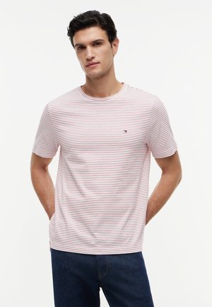 Άνδρας με σκούρα μαλλιά, που φοράει λευκό t-shirt με λεπτές κόκκινες οριζόντιες ρίγες και σκούρο μπλε τζιν, στέκεται μπροστά σε ανοιχτό φόντο.