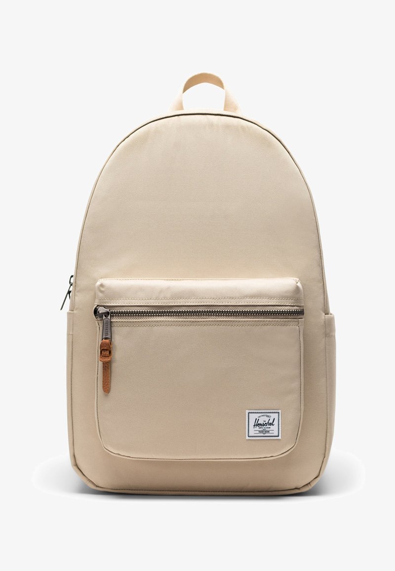 Sac à dos beige Herschel avec poche avant à fermeture éclair et tirette en cuir, étiquette de marque blanche rectangulaire, et poignée de transport supérieure.