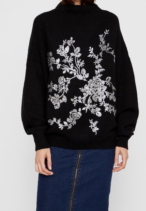 Zwarte gebreide trui met een hoge hals en lange mouwen, voorzien van zilveren bloemenborduring aan de voorkant. Gepaard met een denimrok.
