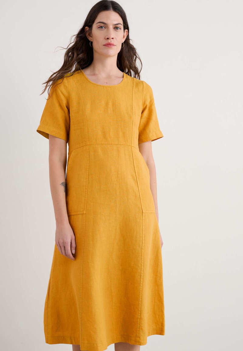 Žluté lněné šaty s krátkým rukávem, kulatým výstřihem, přiléhavým tělem a áčkovým střihem. Obsahují vertikální švy a boční kapsy.