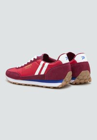 Zapatillas bajas rojas y burdeos con rayas blancas, cordones blancos, suelas medias azules y blancas, y suelas de goma texturizadas, lado a lado sobre un fondo gris.