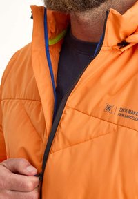 Chaqueta acolchada naranja con cierre frontal de cremallera, forro azul y verde, y detalle de cremallera negra. La tela tiene una apariencia suave y texturada.