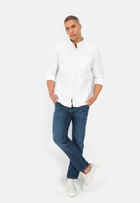 Witte overhemd met lange mouwen en borstzak, gecombineerd met blauwe spijkerbroek en witte sneakers. Staande pose met ontspannen armen.
