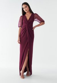 Burgundijas krāsas vakara kleita ar dziļu V veida kakla izgriezumu, savīto priekšpusi, plānām plīvurojošām piedurknēm un augstu šķēlumu. Gluda tekstūra, eleganta siluets.