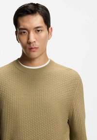 Maglione beige dalla texture strutturata con motivo a griglia, collo tondo a costine e maniche lunghe, indossato da una modella su uno sfondo neutro.
