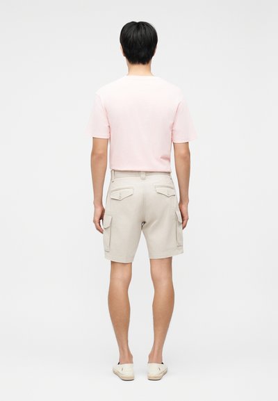 Άνδρας που στέκεται με την πλάτη προς τα εμάς, φοράει ροζ ανοιχτό κοντομάνικο πουκάμισο, μπεζ σορτς με τσέπες και λευκά παπούτσια slip-on, με απλό φόντο.