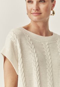 Chunky gebreid sweater in roomwit met korte mouwen, voorzien van kabelbrei-details en een geribbelde halslijn. Glad textuur met een ontspannen pasvorm.