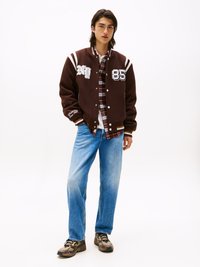 Young man standing wearing brown varsity jacket, red plaid shirt, light blue jeans, and beige sneakers against white background.