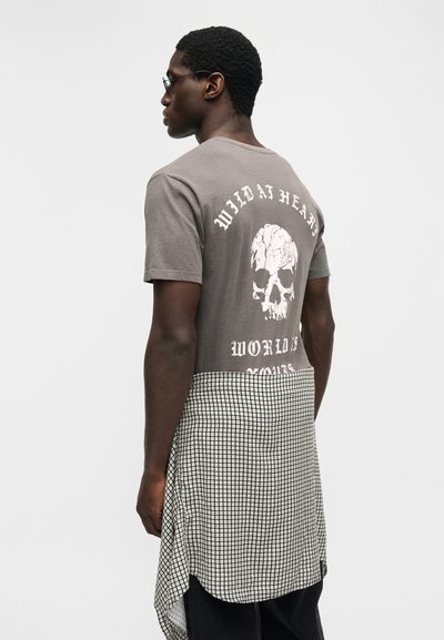 Νεαρός άνδρας με γυαλιά φορώντας γκρι μπλουζάκι με κρανίο και το κείμενο «Wild At Heart», και μια καρό πουκάμισο δεμένο γύρω από τη μέση.