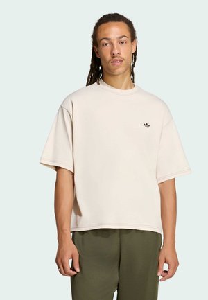 Uomo con lunghi dreadlocks che indossa una maglietta Adidas beige chiaro larga e pantaloni verde scuro su uno sfondo chiaro uniforme.