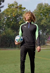 Svart treningsjakke med neongrønne detaljer og kvart-zip, sammen med svarte bukser. Holder en mønstrete fotball på et grønt felt.