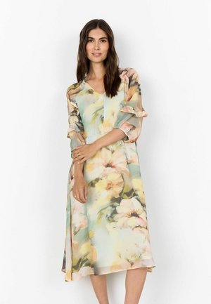 Ženska z dolgimi temnimi lasmi, oblečena v pastelno cvetlično midi obleko z nabrano tročetrtinsko rokavi, stoji pred belim ozadjem.