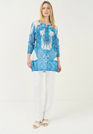 Mujer rubia que lleva una túnica azul y blanca con estampado y mangas largas, pantalones blancos y sandalias abiertas blancas, de pie frente a un fondo liso.