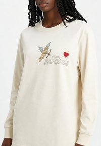 Sweatshirt à manches longues couleur crème avec un graphique brodé d'un chérubin et d'un cœur, accompagné du texte "je t'aime" en police décorative. Matière en coton doux.