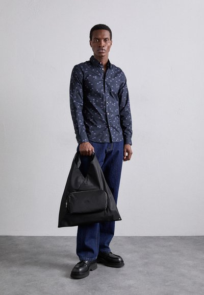Borsa in tessuto nero con forma triangolare, tasca frontale con zip, texture liscia e una larga tracolla. Modello che indossa una camicia a fantasia blu navy e jeans.