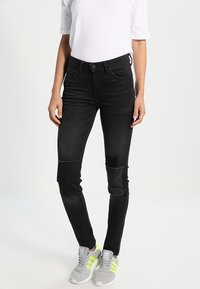 Vaqueros de mezclilla negra con corte slim, que presentan parches en las rodillas, costuras sutiles y una textura suave. Combinados con zapatillas grises y una parte superior blanca.