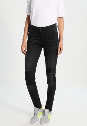 Vaqueros de mezclilla negra con corte slim, que presentan parches en las rodillas, costuras sutiles y una textura suave. Combinados con zapatillas grises y una parte superior blanca.