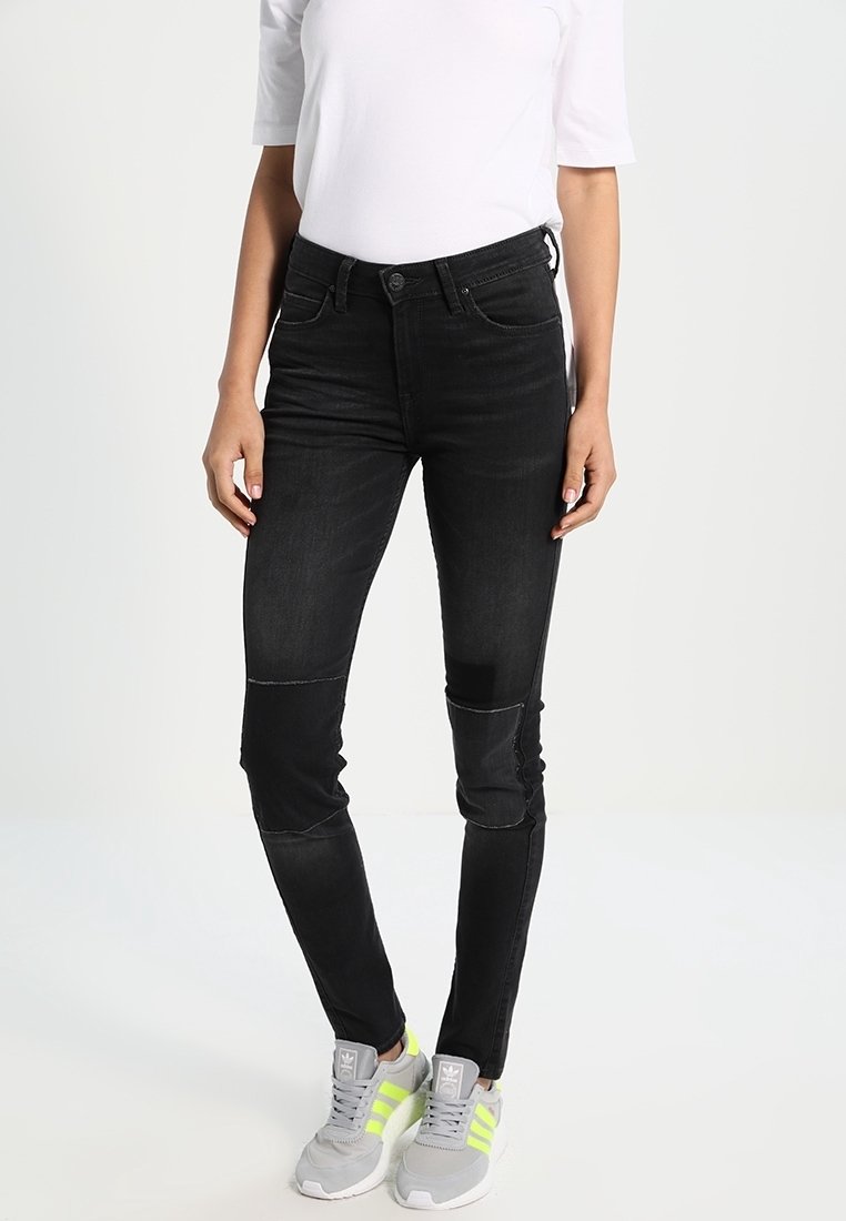 Vaqueros de mezclilla negra con corte slim, que presentan parches en las rodillas, costuras sutiles y una textura suave. Combinados con zapatillas grises y una parte superior blanca.
