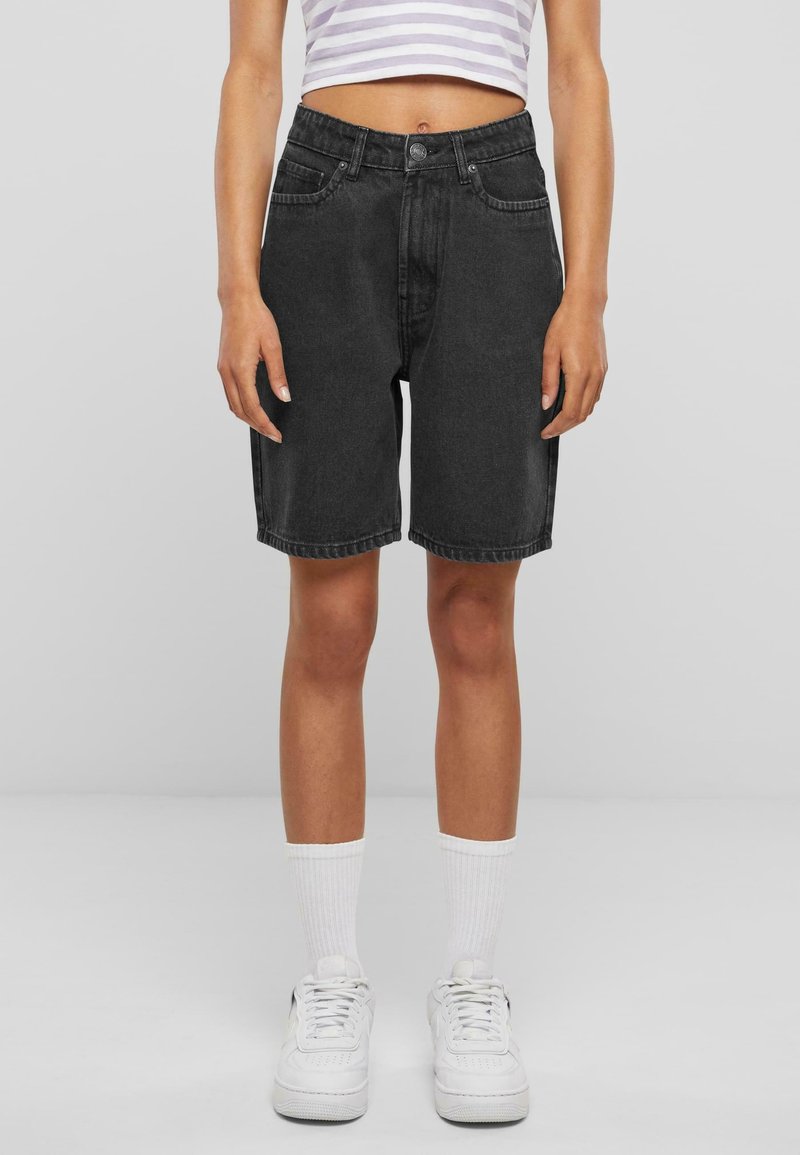 Personne portant un short en denim noir taille haute, des baskets blanches, des chaussettes blanches montantes et un top court blanc rayé gris, debout devant un fond uni.