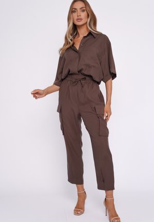 Blonde Frau trägt ein lockeres braunes Hemd mit halblangen Ärmeln und passende Cargohosen, posiert mit einer leicht erhobenen Hand.