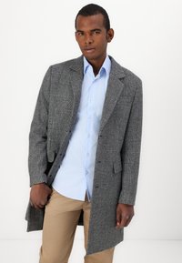 Grau karierten Übermantel mit Revers, getragen über einem hellblauen Hemd. Das Modell hat dunkle Haut und steht vor einem weißen Hintergrund.