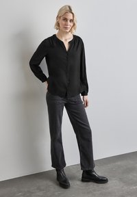 Blusa nera con bottone, scollatura tonda, maniche lunghe e dettagli arricciati sulle spalle, abbinata a jeans grigi scuri e stivali neri.